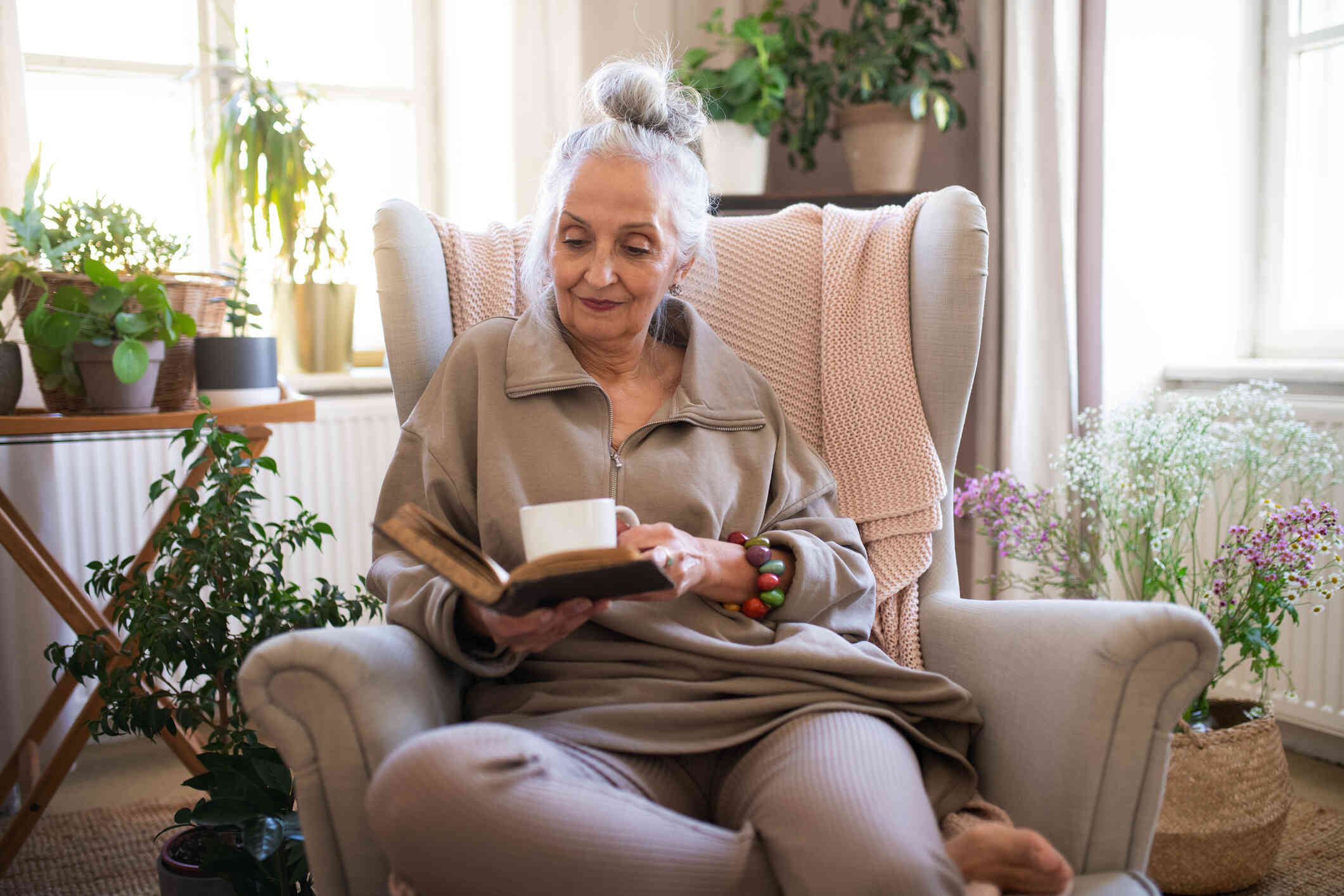 A mature woman sits in an armchair in her home with a cup of coffee and holds her journal open in the other hand.